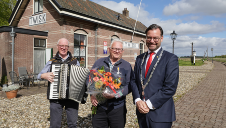 Jan Jong staat naast burgemeester Michiel Pijl aan het station in Medemblik. Meneer de Jong heeft een groot boeket oranje bloemen in zijn handen en hij draagt het lintje.